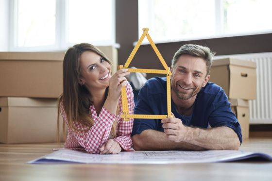 Un couple tient un mètre d'architecte entre les mains qui représentent une maison. Ils sont allongés au sol sur des papiers qui contiennent les côtes de leur maison. Derrière eux, des cartons de déménagement.