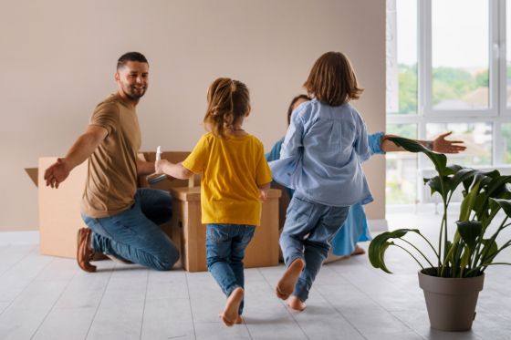 Deux enfants courent vers leurs parents dans un salon. Il y a des cartons dans le salon.